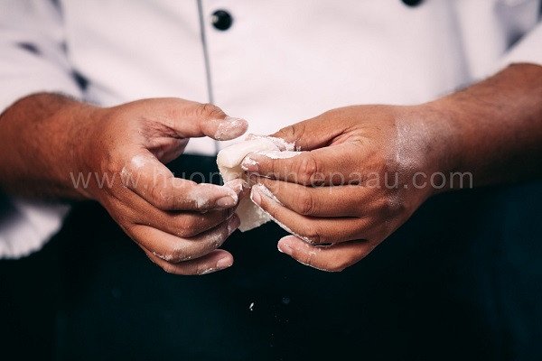Bàn tay đang nhào bột mochi mềm dẻo trên thớt gỗ, xung quanh có các dụng cụ làm bánh và nguyên liệu