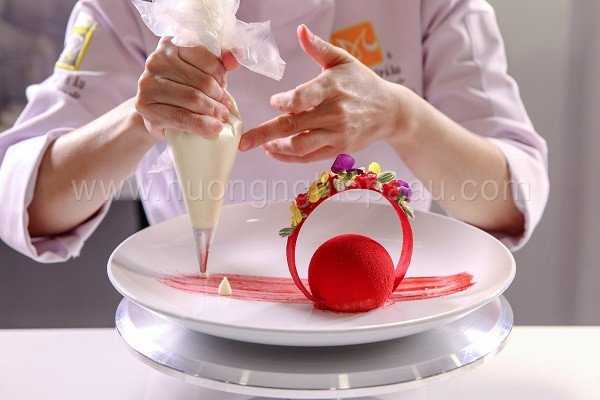 Người thợ bánh đang hoàn thiện lớp trang trí gương sáng bóng cho chiếc bánh Entremet sô cô la tinh xảo, thể hiện kỹ thuật làm bánh chuyên nghiệp và tỉ mỉ.