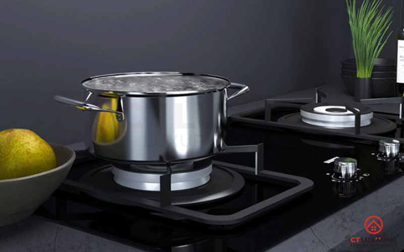 Một người phụ nữ đang vệ sinh đầu đốt của bếp ga bằng bàn chải nhỏ, phía sau là nồi cháo.